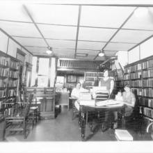 looking E from inside the library when it was located in the Times Newspaper Office, 132 N Main St.; handwriting on the back reads, "Bertha Spicer at back desk 1927" "Dawn Spicer, dau. of Bertha, standing beside her." "Mrs. Roy Hays + Little Sarah Hays at table." "Pardeeville Library located in building on east side of No. Main St. where Steve Thompson's Printing Office is now (1992)."